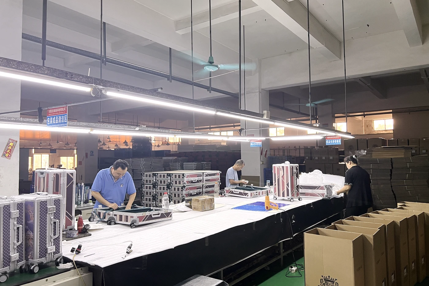 Workers assembling luggage at factory workbenches with protective packing materials and cartons nearby