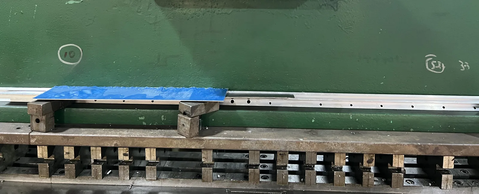 Aluminum luggage frame extrusion resting on the die bed of a double-sided hydraulic punch, with the punched hole row visible along the profile