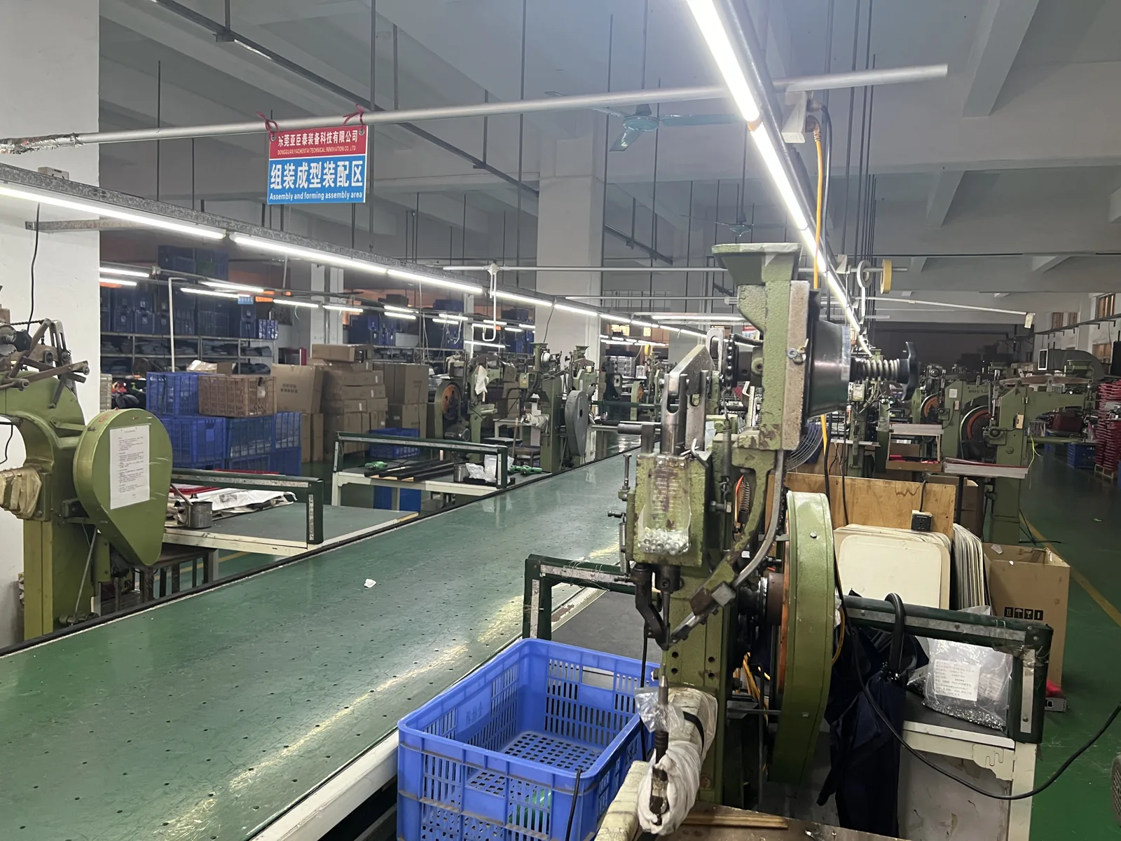 Assembly area with presses and conveyor, blue crates of components and stacked cartons along the aisle