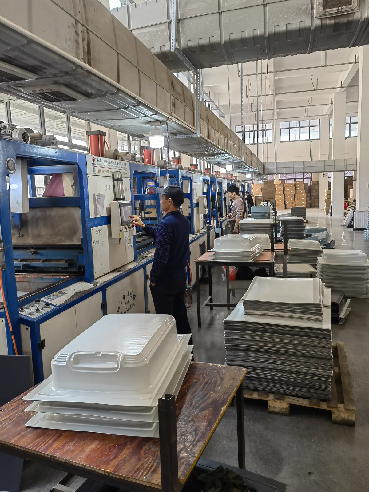 Line of thermoforming presses shaping luggage shells from heated plastic sheet, with cut sheet stock stacked alongside